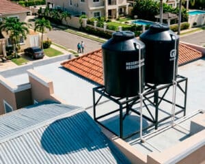 Vista en primer plano desde una azotea donde destacan dos grandes tinacos negros sobre una estructura metálica. A la izquierda, los tinacos proyectan una sombra pronunciada y ovalada sobre un techo de zinc corrugado. De fondo, se observa una calle de un sector residencial tranquilo, con casas de dos niveles, techos de tejas, palmeras, un vehículo negro estacionado y dos personas caminando por la acera.