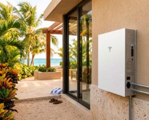 Batería de pared blanca Tesla montada en la pared exterior de estuco de una casa moderna de estilo tropical frente al mar en un día soleado. A la izquierda, exuberantes palmeras, plantas de crotón de colores y arena de playa con sandalias conducen a una vista al océano azul turquesa. La casa tiene una gran puerta corrediza de vidrio.